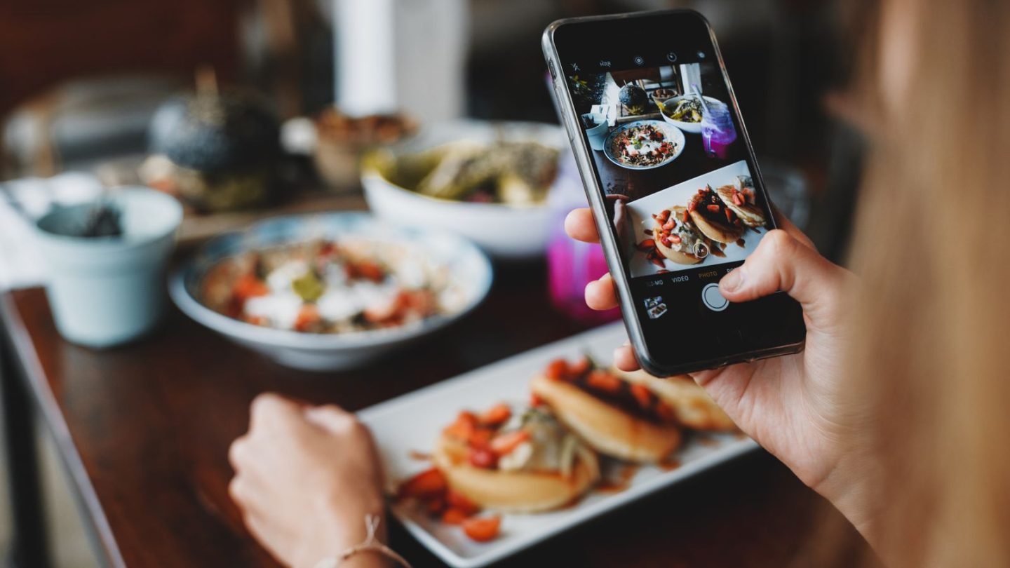 Hand holding a smartphone to photograph plates of food on a cafe table
