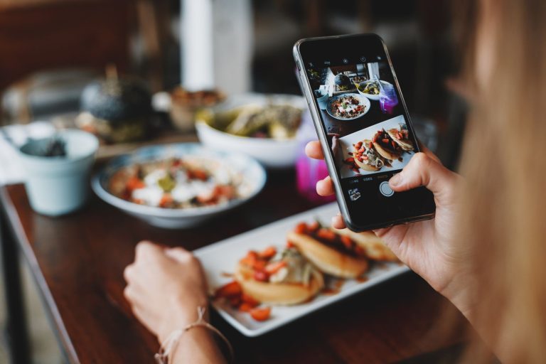 Hand holding a smartphone to photograph plates of food on a cafe table