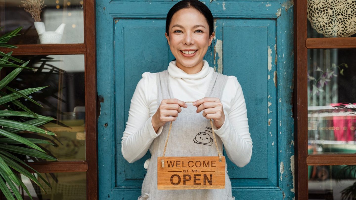 Smiling small business owner in an apron holding a Welcome We Are Open sign in front of her shop door