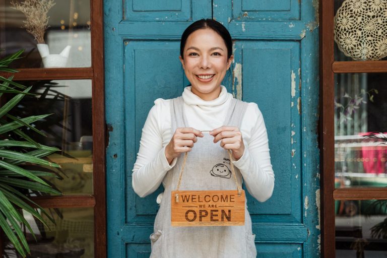 Smiling small business owner in an apron holding a Welcome We Are Open sign in front of her shop door