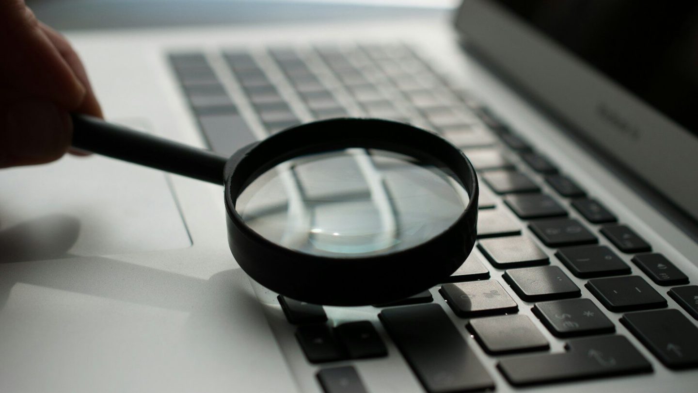 Hand holding a magnifying glass over a laptop keyboard, representing a WordPress speed test