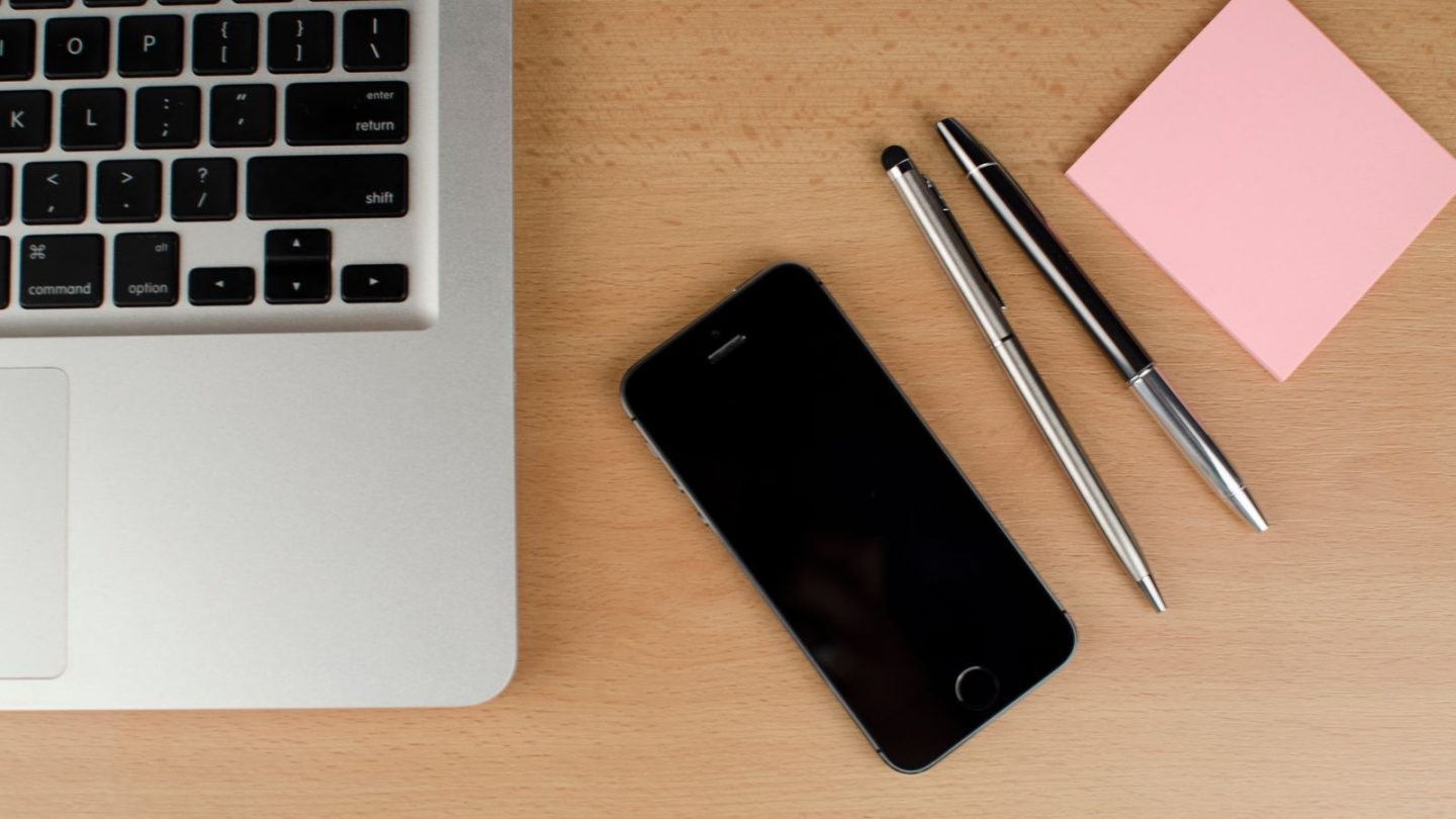 Smartphone, laptop keyboard, pens and a sticky note on a wooden desk