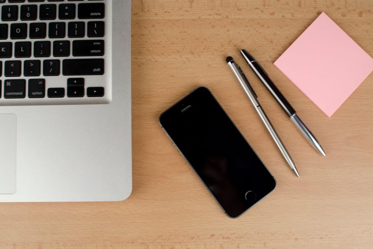 Smartphone, laptop keyboard, pens and a sticky note on a wooden desk