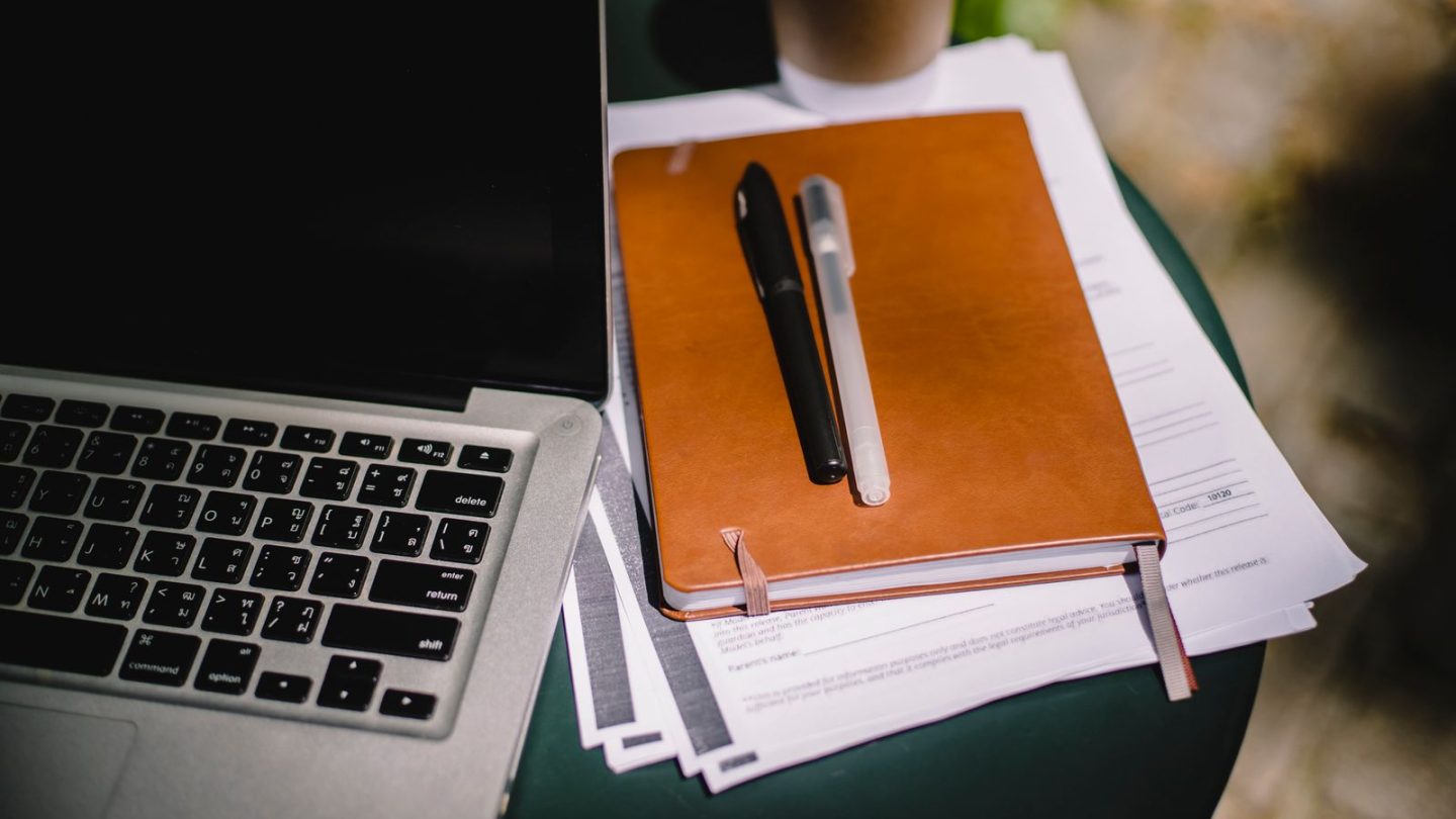 Laptop beside a leather notebook and papers on an outdoor table