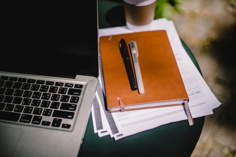 Laptop beside a leather notebook and papers on an outdoor table